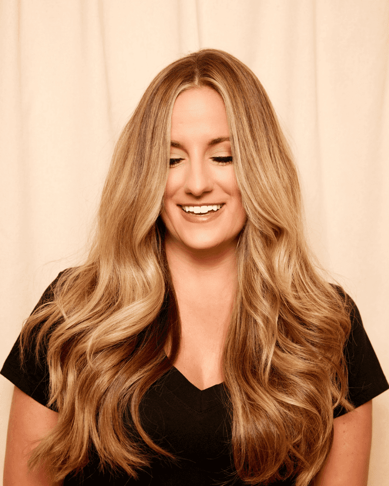 Smiling woman with long blonde wavy hair, wearing a black top, against a beige background.